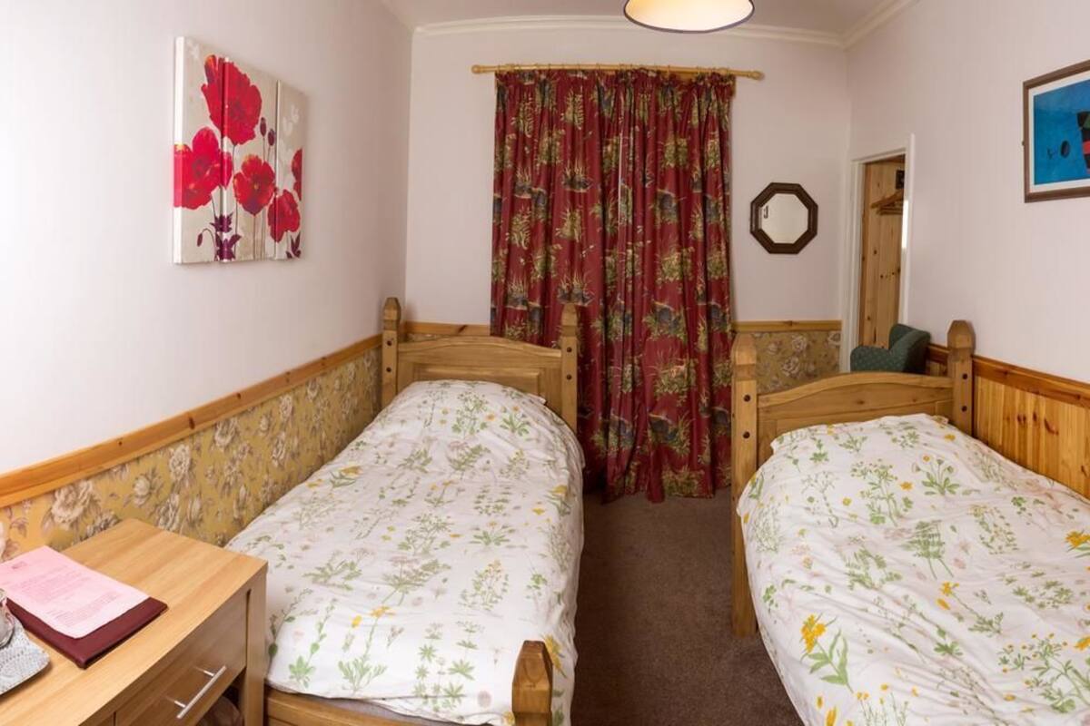 Quarto com duas camas de madeira com roupa de cama florida, uma escrivaninha de madeira com cadeira, um espelho e um quadro na parede.