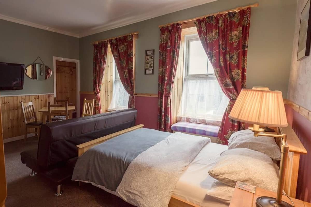 Quarto com cama, mesa de cabeceira, abajur e janela com cortinas.