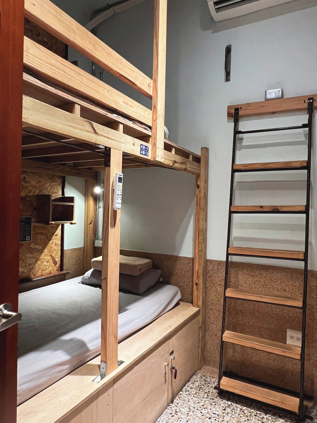 A bunk bed room with a wooden ladder, a small desk, and a wall-mounted air conditioning unit.