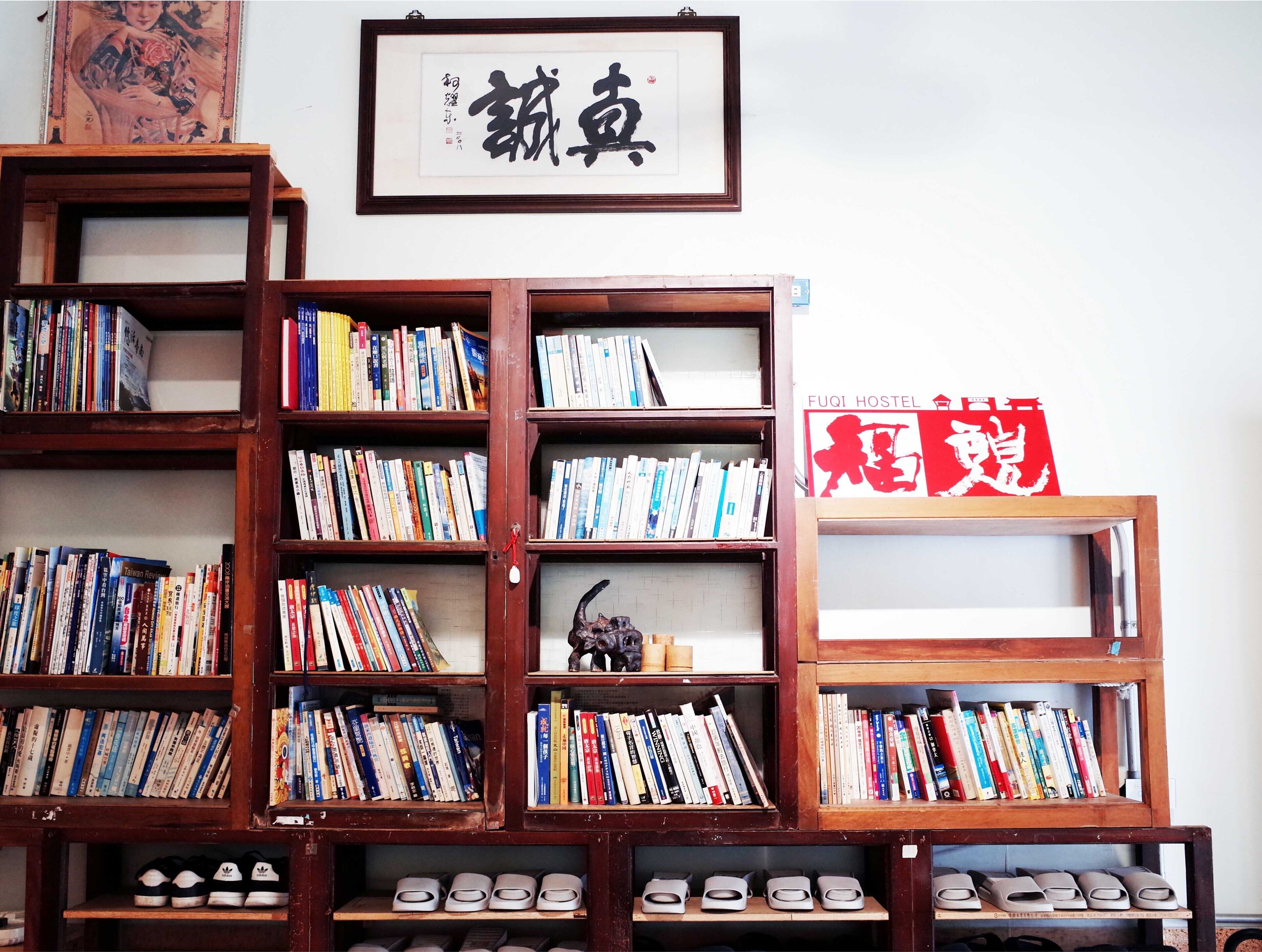 A room with wooden bookshelves filled with books, a framed calligraphy piece, and a sign for FUQI HOSTEL.