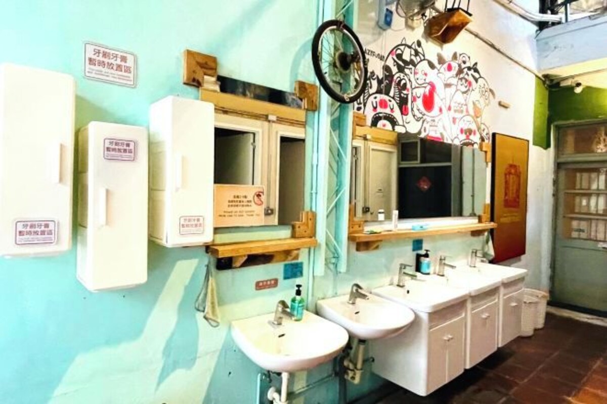 A bathroom with two sinks, a mirror, and a toilet. There are signs on the wall and a shelf with toiletries.