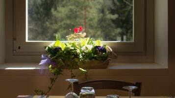 Une table à manger avec vue sur une forêt par la fenêtre, un vase rempli de fleurs et une bougie.