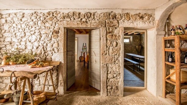 A rustic stone-walled room with a wooden table, benches, and a wine rack.