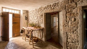 A rustic stone-walled room with a wooden table and chairs, a large open door, and a stone archway.