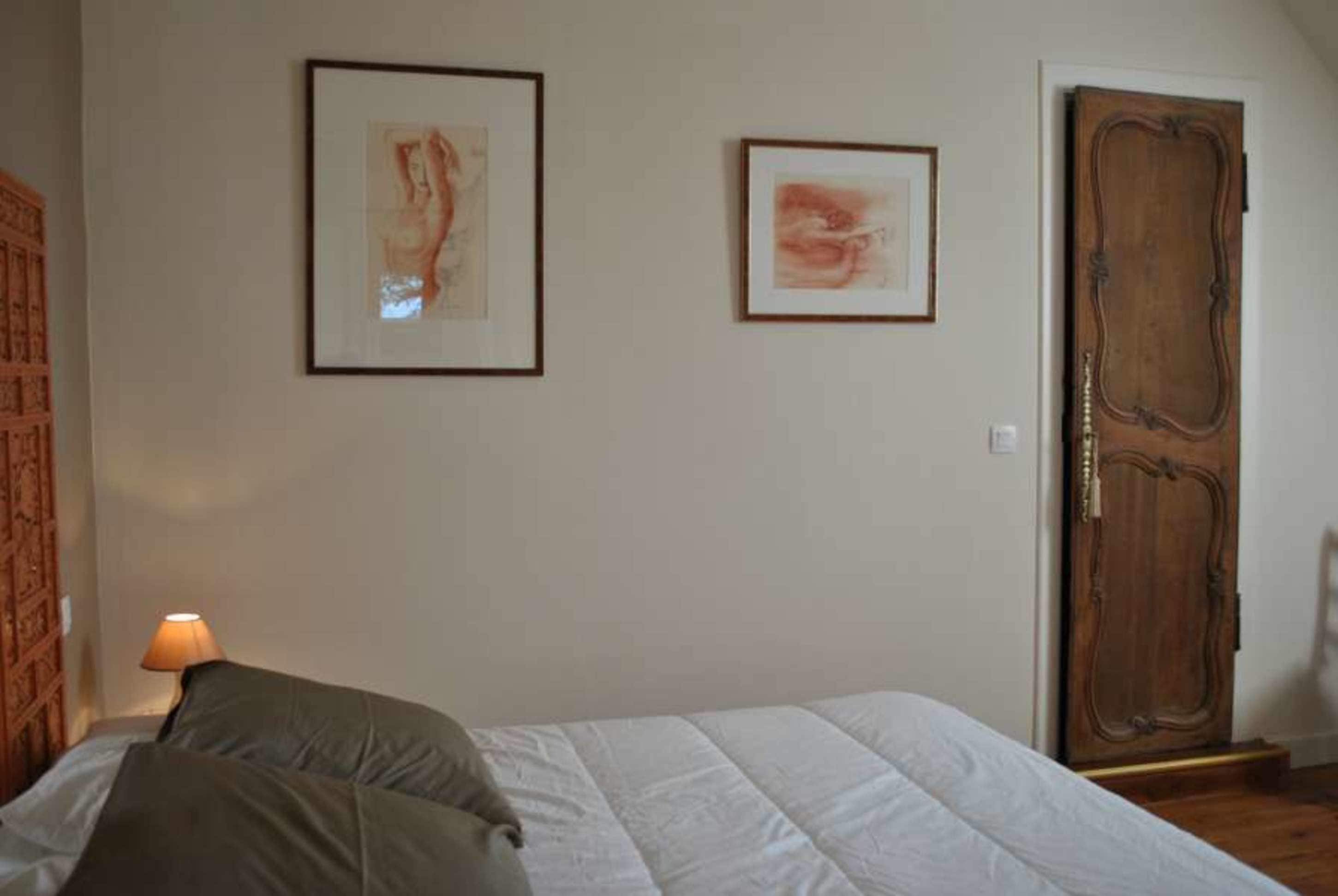 Un dormitorio con cama, dos cuadros enmarcados en la pared, una puerta de madera y una lámpara en una mesita de noche.
