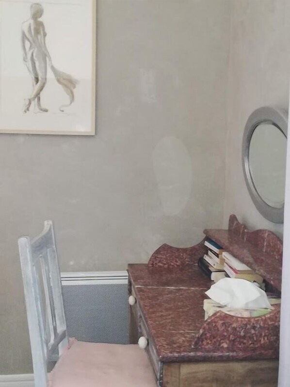 Una habitación con cama, una silla, un escritorio con libros, un espejo y un cuadro en la pared.