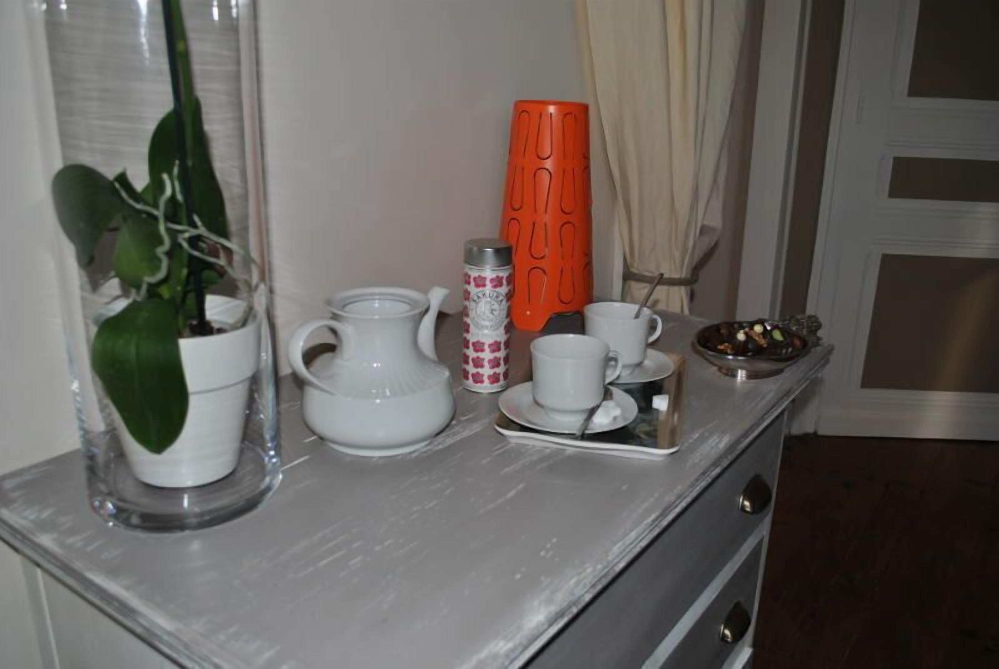 Uma bancada de cozinha com uma planta em vaso, um jarro branco, uma garrafa térmica, uma caneca, uma xícara e uma bandeja com uma colher.
