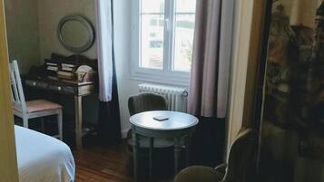 A small, cozy room with a bed, a chair, a table, and a window with curtains.
