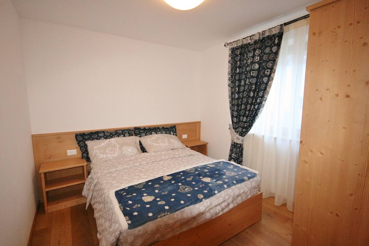 Une chambre à coucher avec un lit en bois, une table de chevet, une armoire en bois et une fenêtre ornée de rideaux à motifs.