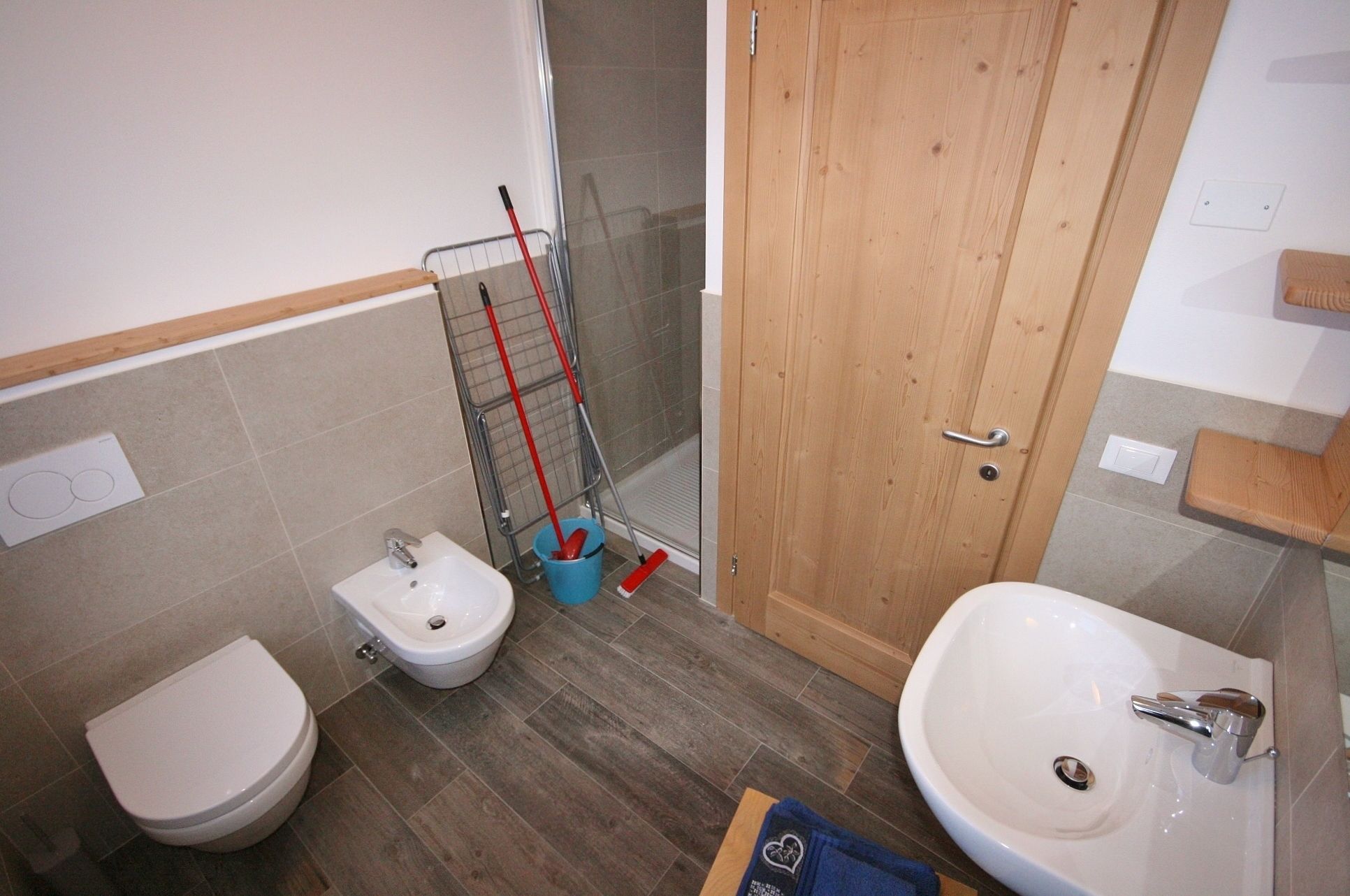 A bathroom with a toilet, bidet, and sink. There is a wooden door, a shower area, and a cleaning cart with mops.