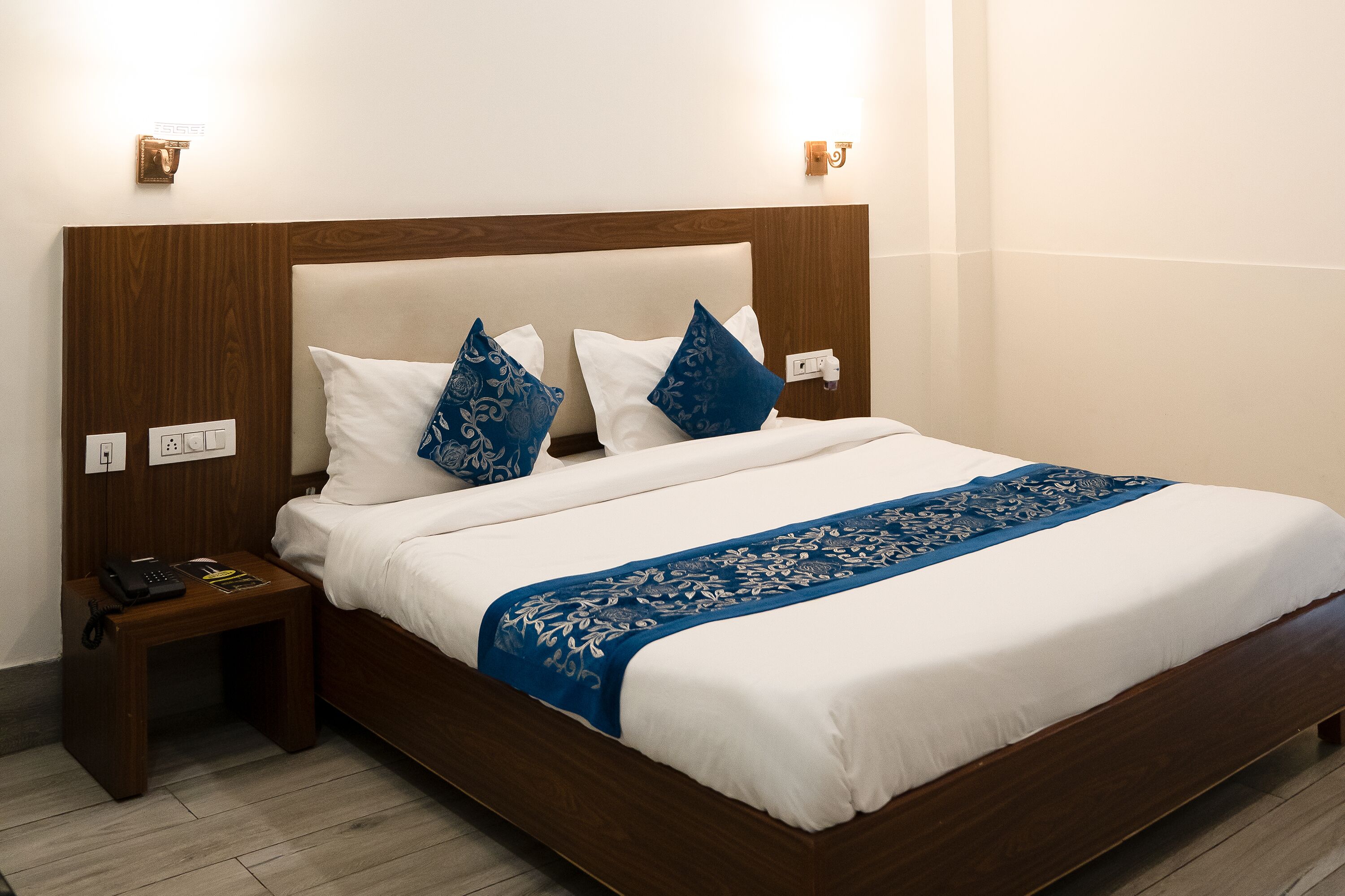 Una cama bien hecha con cabecera de madera, almohadas estampadas en blanco y azul, y una manta bordada en azul.