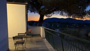 Una terraza al aire libre con una mesa y sillas, una barandilla y vistas a una cadena montañosa al atardecer.