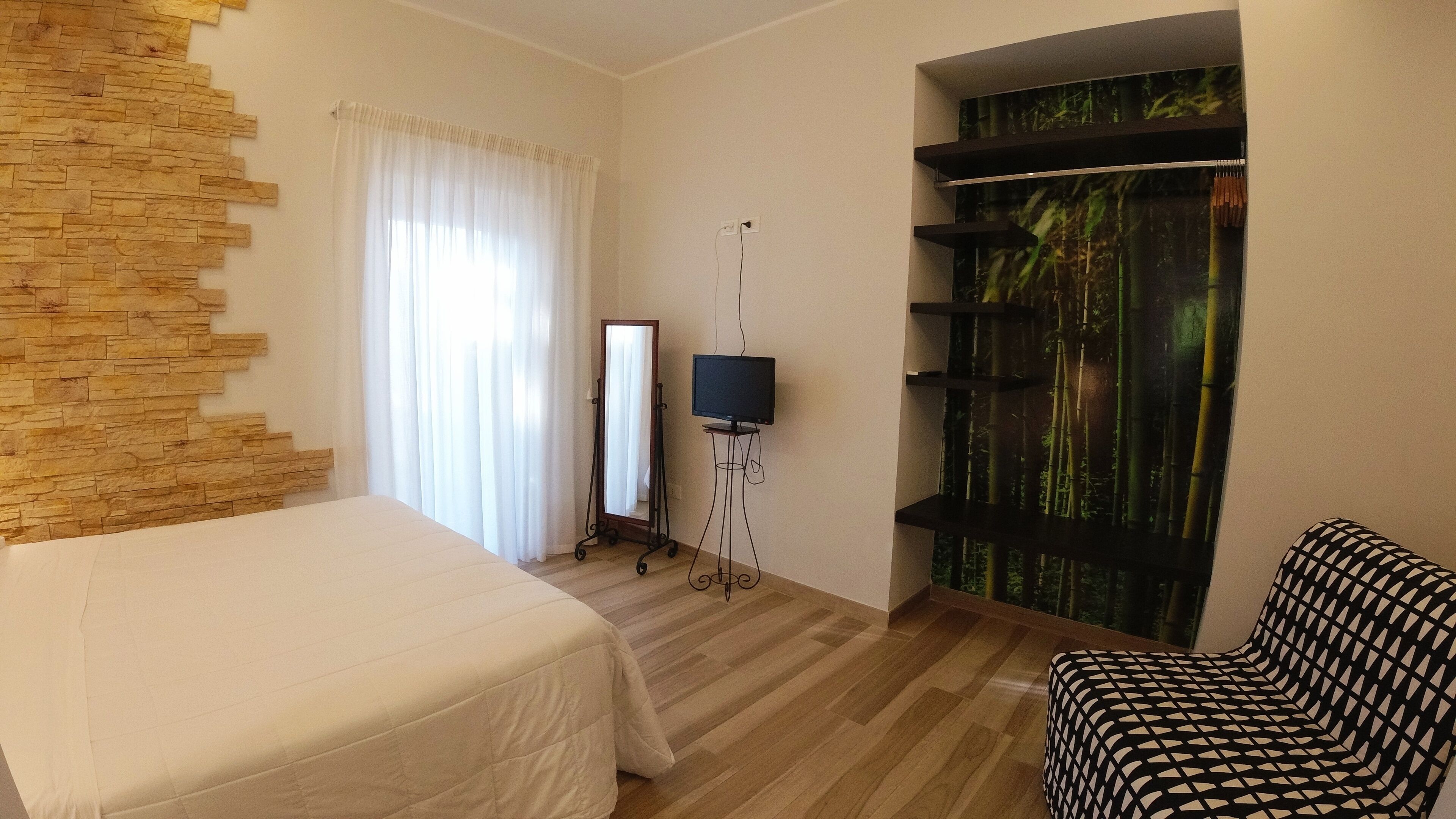 Habitación de hotel con una cama, una silla, un televisor sobre un soporte, un espejo y un cuadro de bambú en la pared.