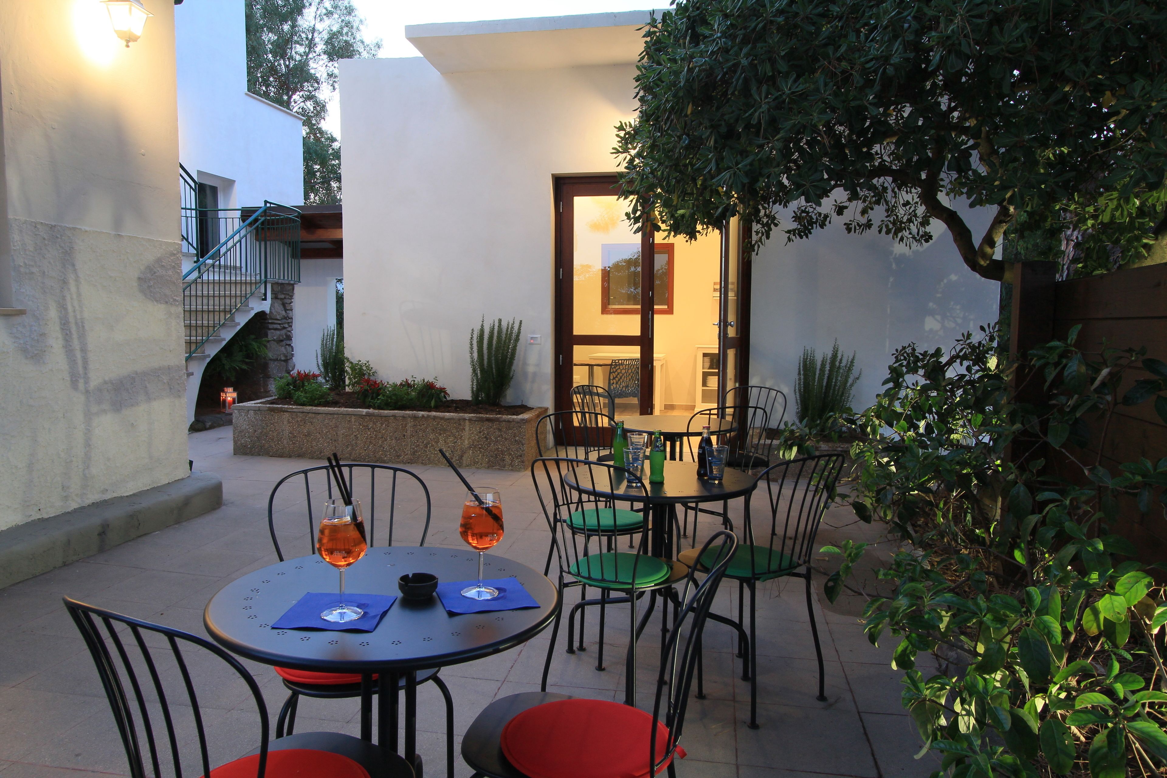 Un patio al aire libre con mesas y sillas redondas, una escalera y vista del interior a través de una puerta de cristal.