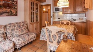 A cozy living room with a floral sofa, a wooden dining table with chairs, and a kitchen area with wooden cabinets and a hanging light fixture.