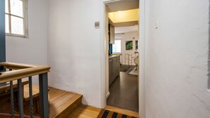 Een smalle gang met een houten trap die leidt naar een kamer. Een deur opent naar een keuken met moderne apparatuur en uitzicht naar buiten.