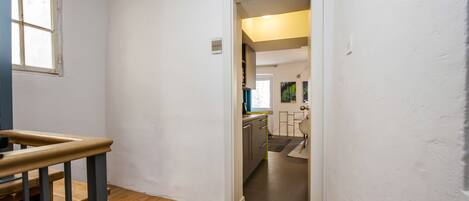 A narrow hallway with a wooden staircase leading to a room. A door opens to a kitchen area with modern appliances and a view of the outside.