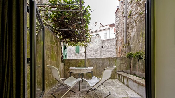 Een klein terras buiten met een tafel en twee stoelen, een stenen vloer en zicht op een stenen muur en gebouwen op de achtergrond.