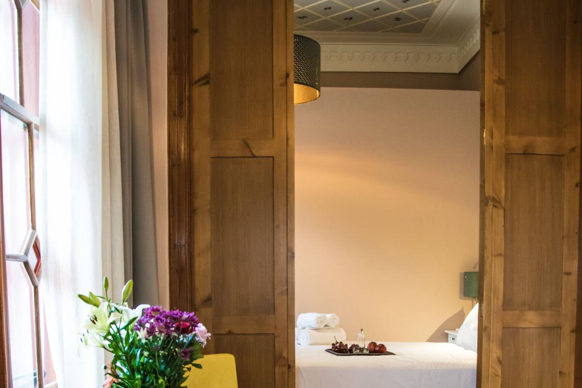 Ein Zimmer mit einem Bett, einem Sessel, einem kleinen Tisch mit Blumenvase und einem Fenster mit Vorhängen.