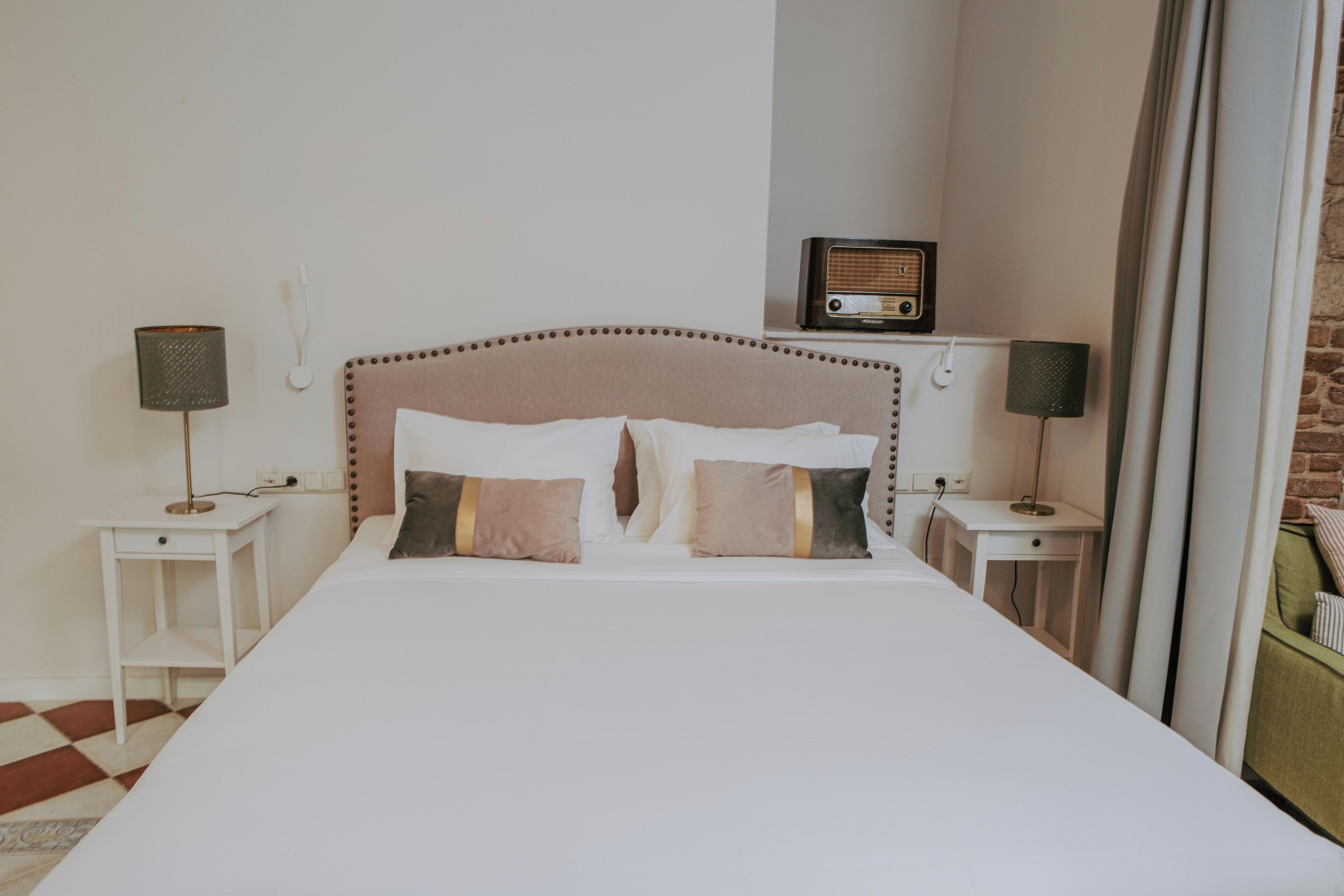 A neatly made bed with a headboard, two bedside tables with lamps, a radio, and a brick wall.
