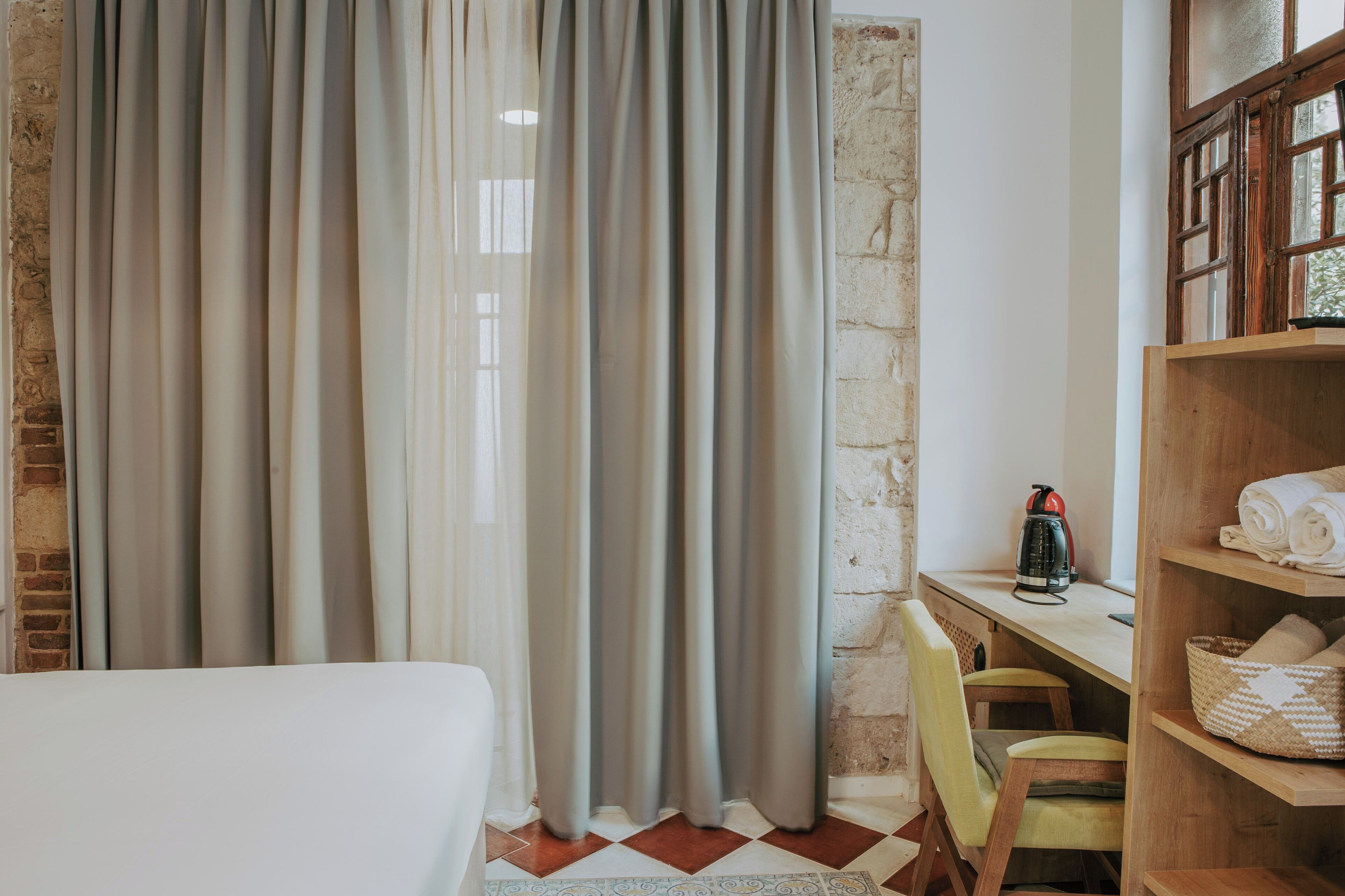A hotel room with a bed, a desk with a kettle, a chair, and a shelf with towels.