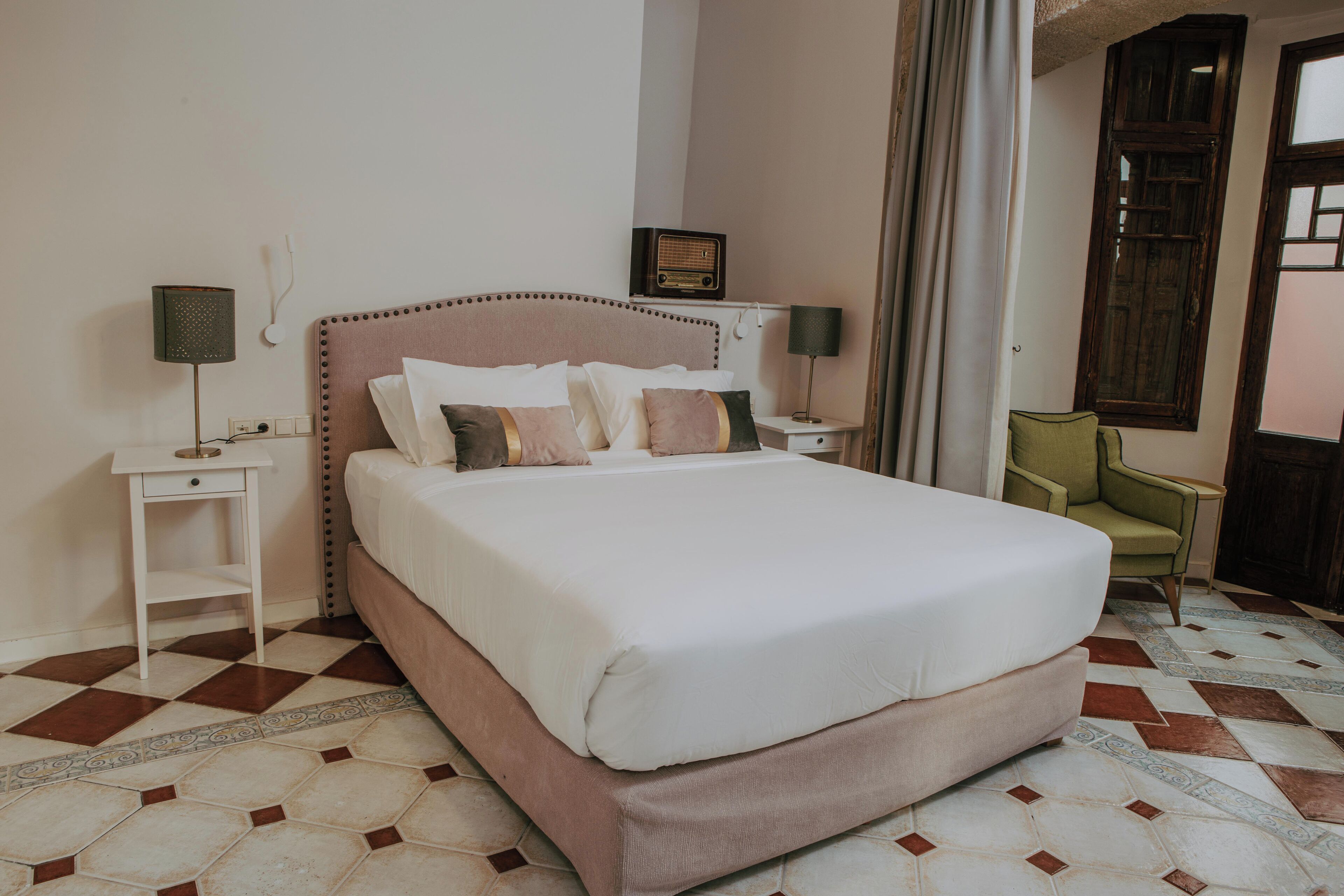 Una cama bien tendida en una habitación de hotel, con un suelo a cuadros, una mesita de noche con una lámpara y una silla.