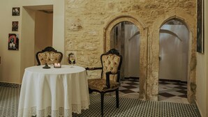 A dining area with a round table, two chairs, and a candle. There are two arched doorways leading to another room. The walls are made of stone, and the floor has a checkered pattern.