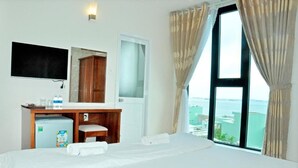 A hotel room with a bed, a desk with a mirror, a small refrigerator, and a window with curtains. - Tram Anh Hotel (Quy Nhon)
