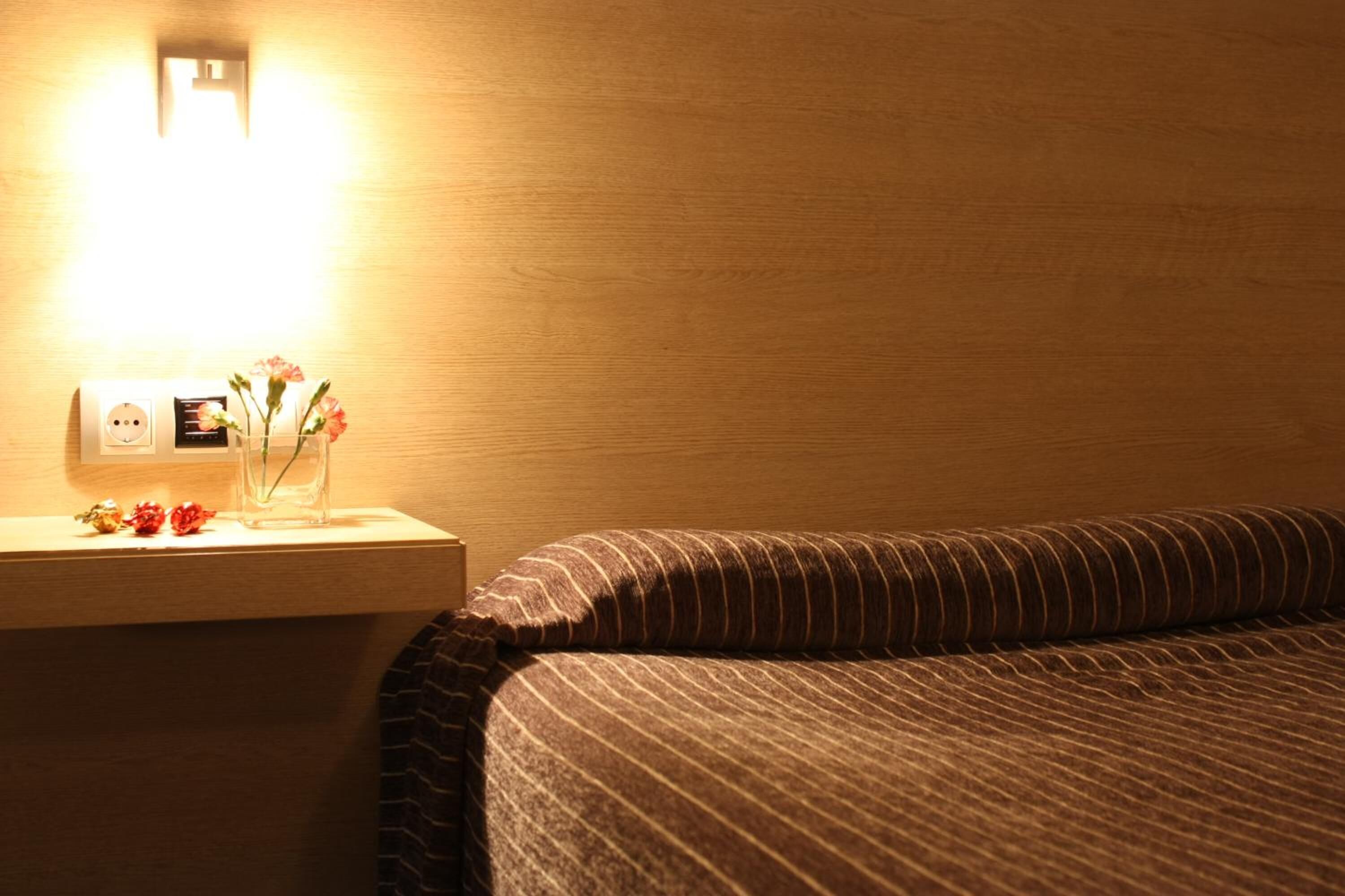 A hotel room with a wooden headboard, a bedside table with a lamp, and a nightstand with a vase of flowers.