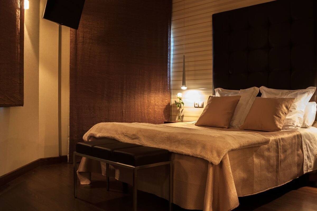 Un dormitorio con cama, banco, mesita de noche y una lámpara de pared.