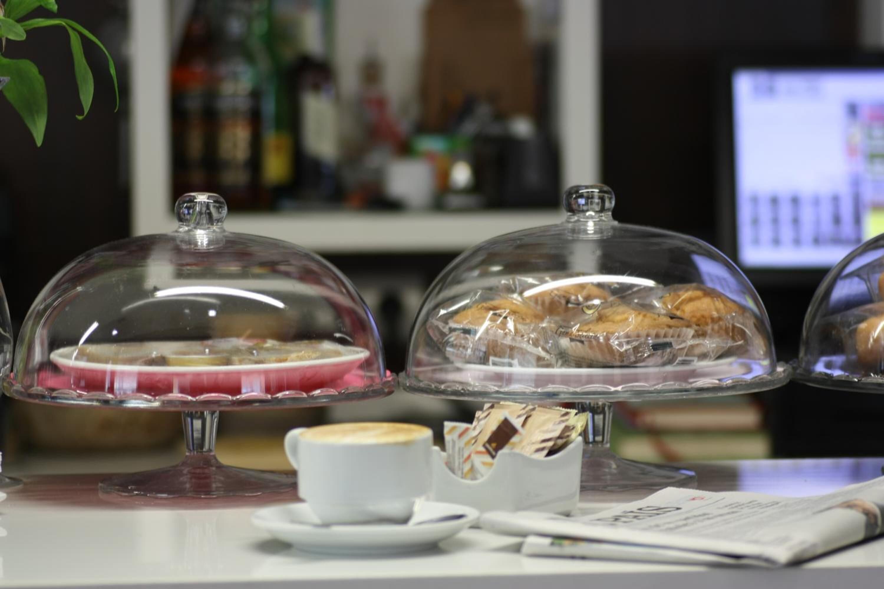 A breakfast setup with pastries under glass domes, a coffee cup, and a newspaper on a table.