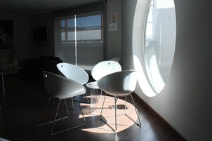 Quarto moderno com três cadeiras brancas, uma mesa pequena e uma janela grande que oferece luz natural.
