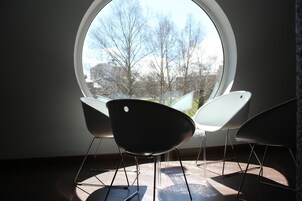 Quarto com uma grande janela redonda, duas cadeiras modernas e vista para árvores e edifícios.