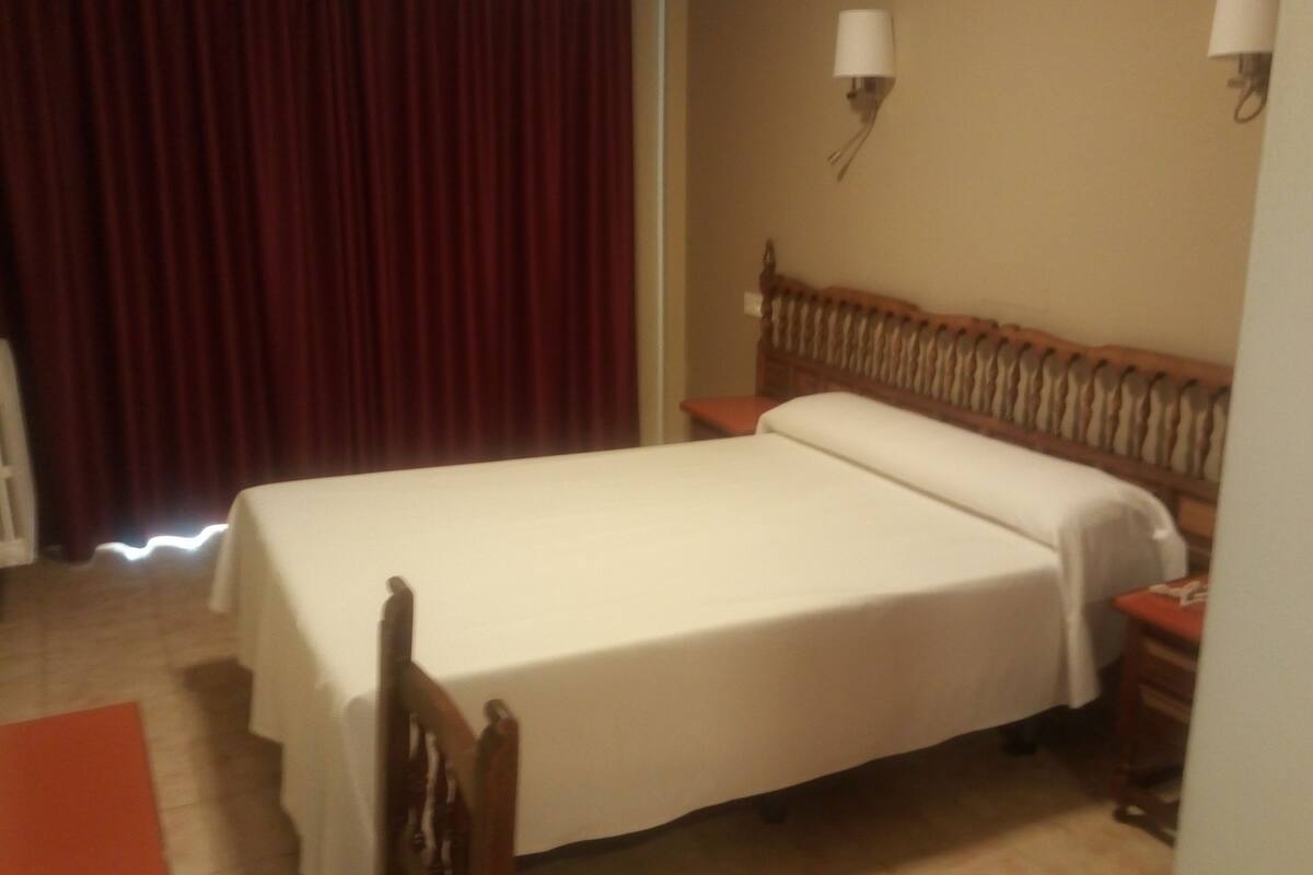 Habitació d'hotel amb un llit, una tauleta de nit, una làmpada i una cortina.