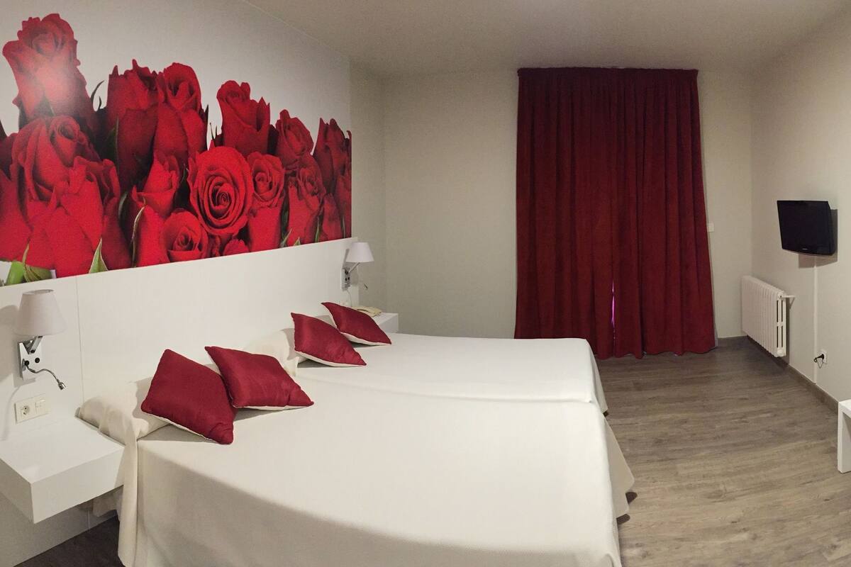 Una habitació d'hotel amb dos llits, una taula petita, una panxa, una televisió i un mural gran amb roses vermelles.