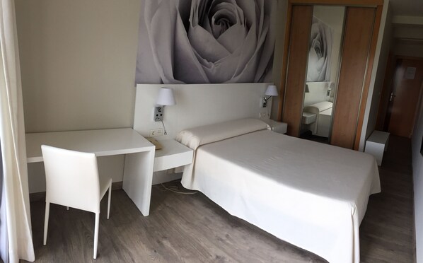 Una habitación de hotel con cama, escritorio y silla. Hay un espejo grande en la pared y un cuadro de una rosa.