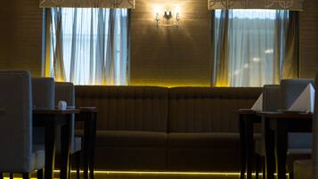 Una habitación con poca luz, un sofá y dos sillones, una ventana con cortinas, y una lámpara en la pared.