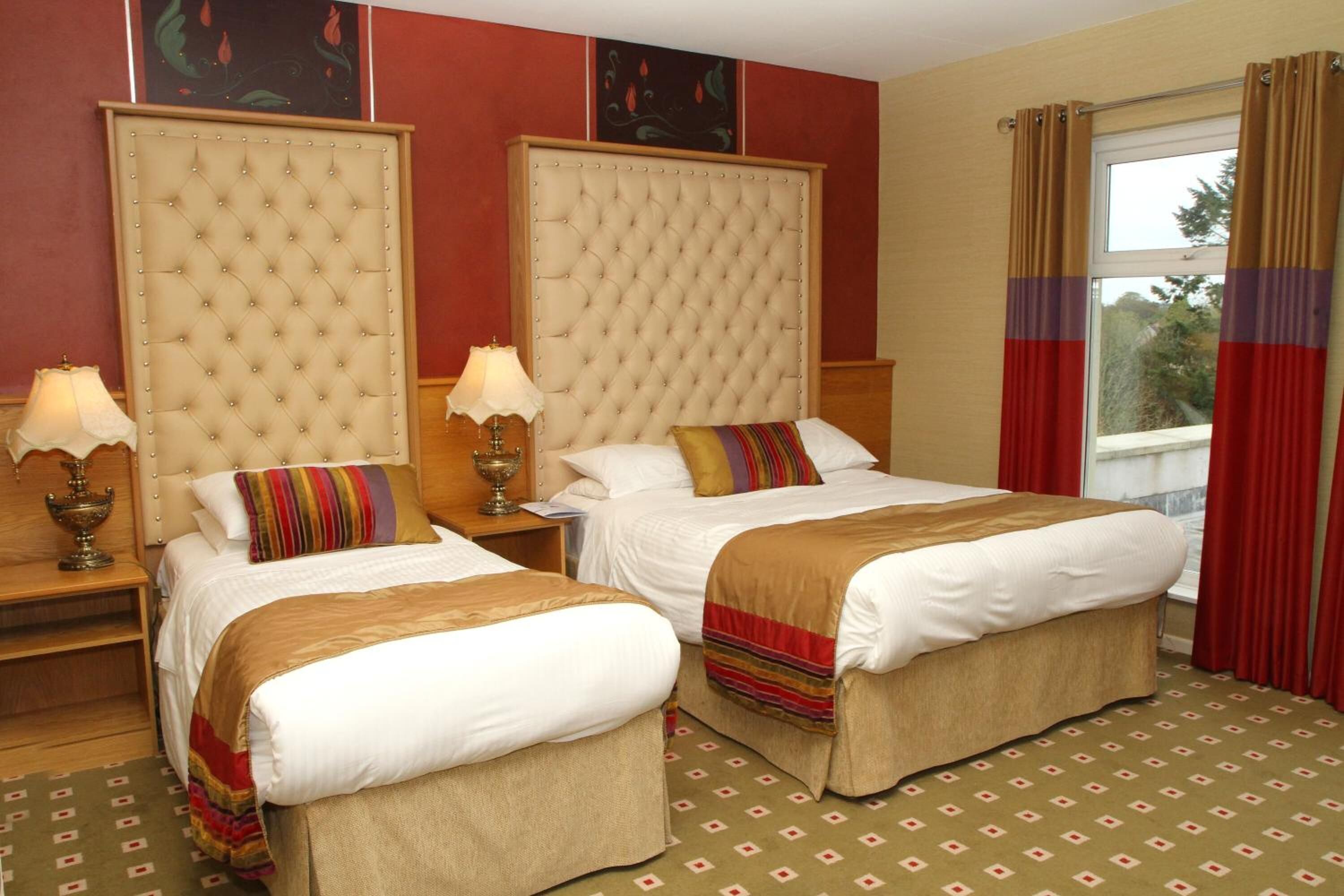 Habitación de hotel con dos camas, un cabecero grande y acolchado, una ventana con cortinas rojas y una alfombra estampada.