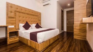 Une chambre d’hôtel moderne avec une tête de lit en bois, un lit recouvert de draps blancs et agrémenté d’oreillers violets, un plancher en bois et un téléviseur à écran plat fixé au mur.
