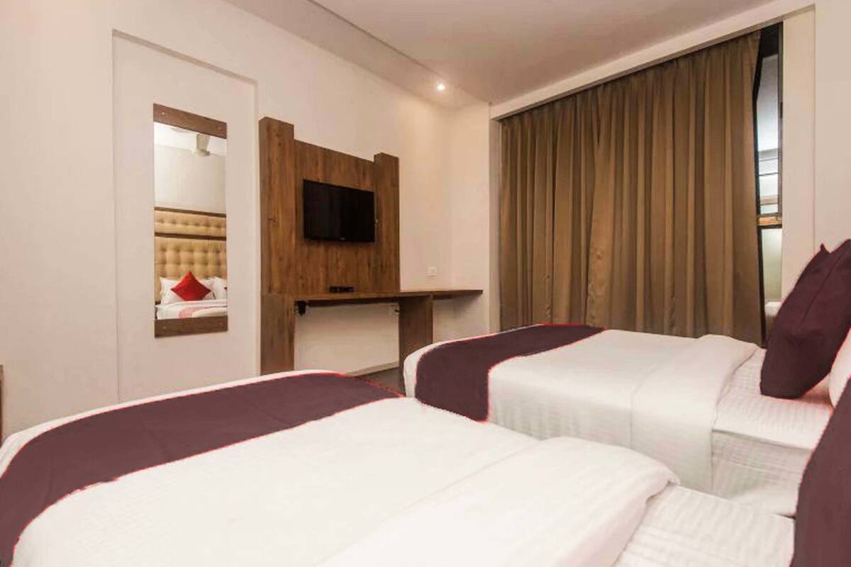 Habitación de hotel con dos camas, un televisor instalado en un panel de madera y un ventanal con cortinas.