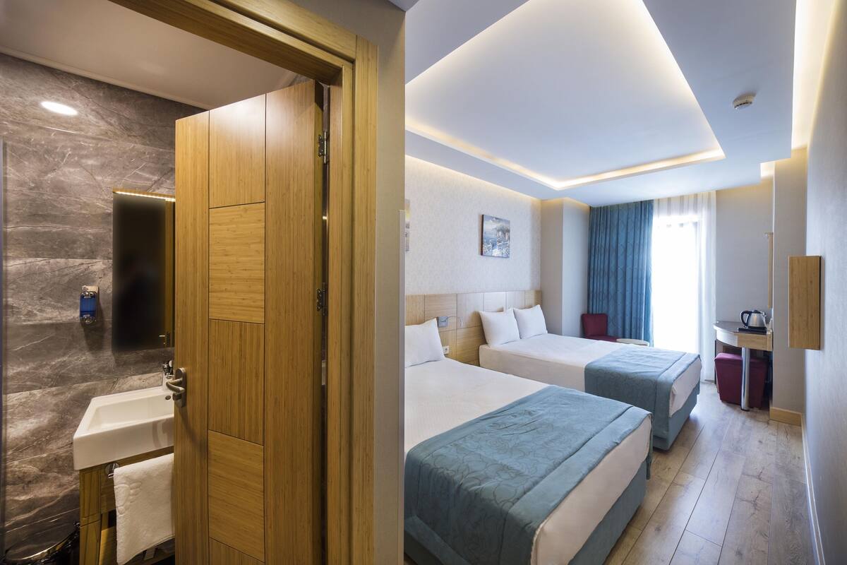 A hotel room with a wooden door, a bathroom with a sink, and two beds with blue bedding.