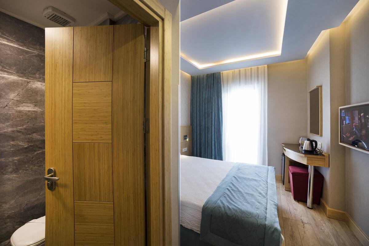 A hotel room with a wooden door, a bed with a blue bedspread, a desk with a chair, a television, and a window with curtains.