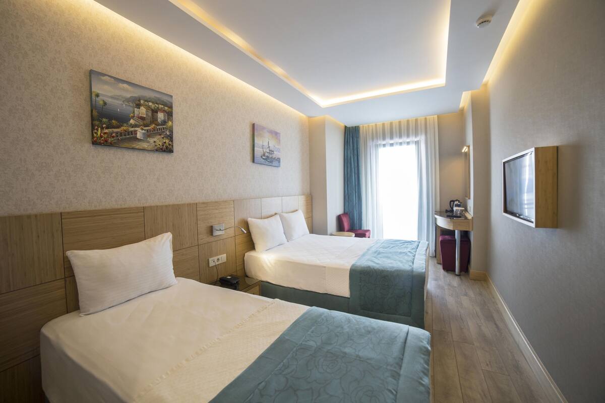 A hotel room with two beds, a desk, a chair, a window with curtains, and artwork on the walls.