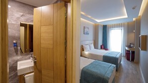 A hotel room with a wooden wardrobe, a bed, a bedside table, a mirror, a sink, and a window with curtains.