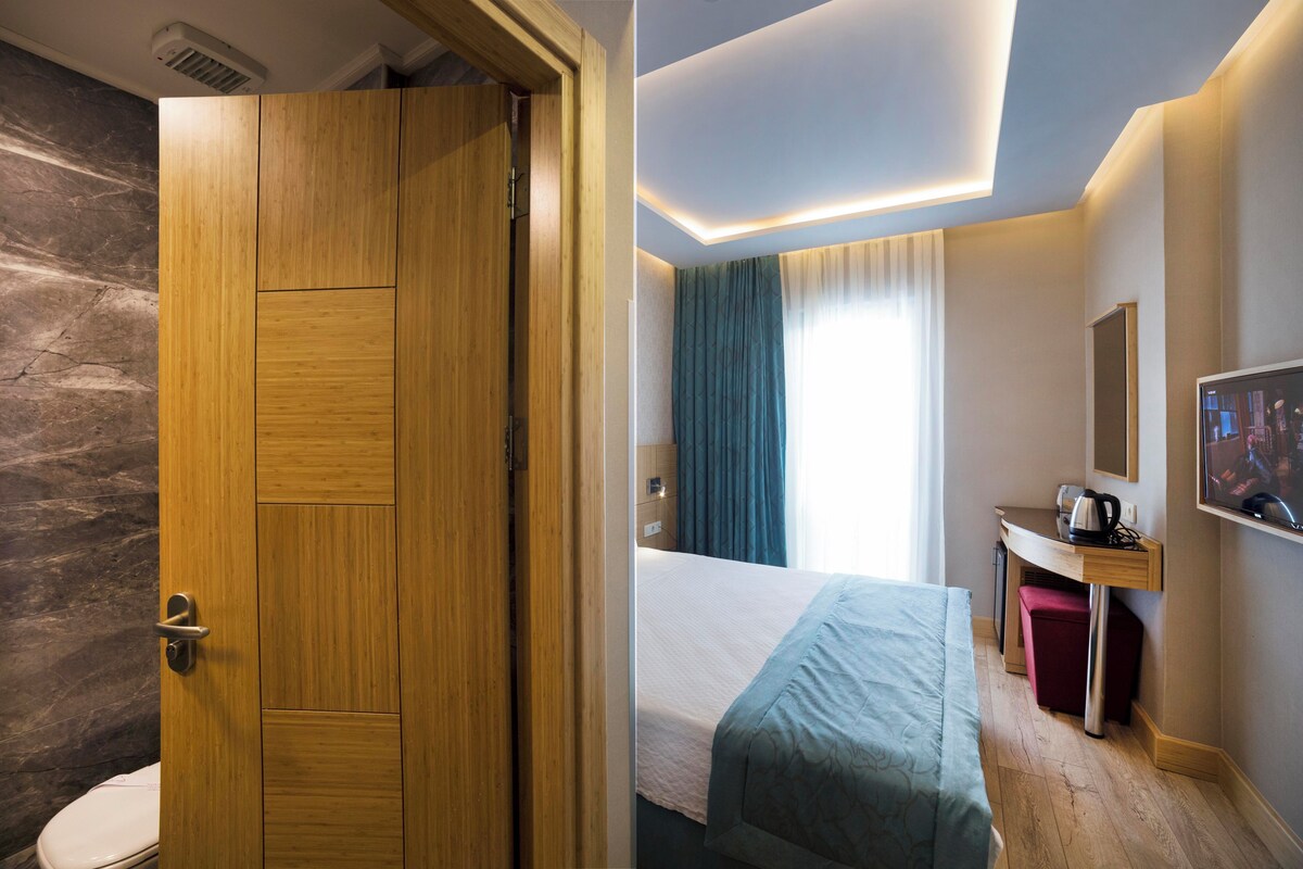 A hotel room with a wooden door, a bed, a desk, a mirror, and a television.