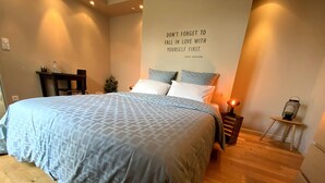 Ein ordentlich bezogenes Bett mit einem gemusterten Überwurf, ein Nachttisch mit Lampe und ein Wandspruch.