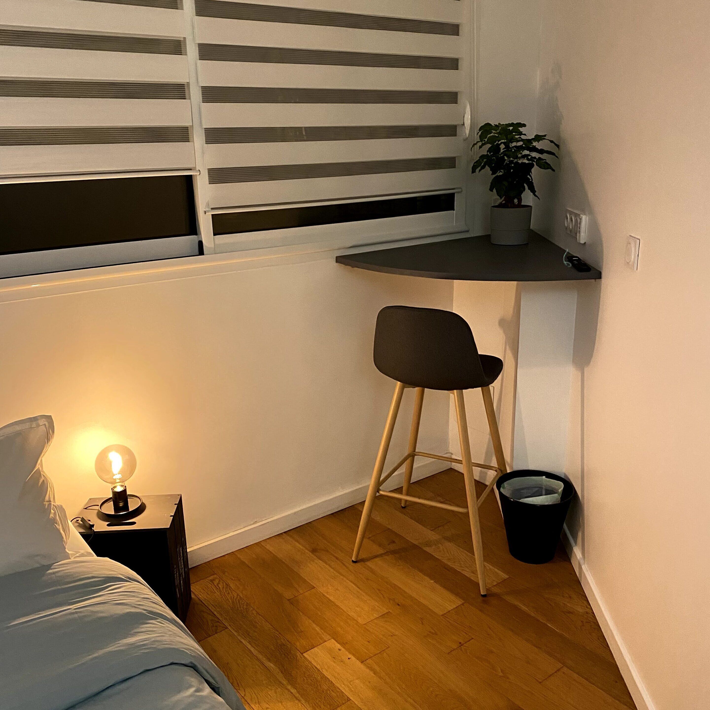 A small, cozy room with a bed, a desk with a chair, a potted plant, and a nightstand with a lamp.