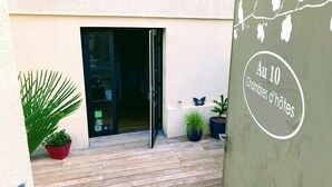 Eine Holzterrasse mit Blumentöpfen, ein Glastür-Eingang und eine Wand mit einem runden Schild, auf dem 'Au 10 Chambres d'hôtes' steht.