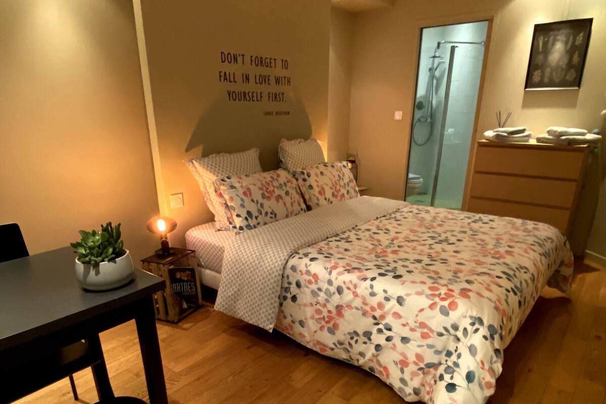 A bedroom with a bed, a bedside table with a lamp, a small plant, and a wall with a motivational quote.