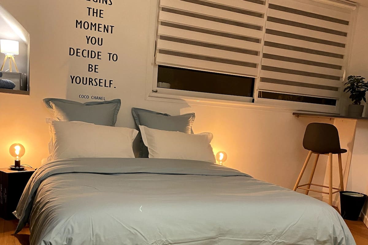 A bedroom with a bed, bedside lamps, a wall with a motivational quote, and a window with blinds.